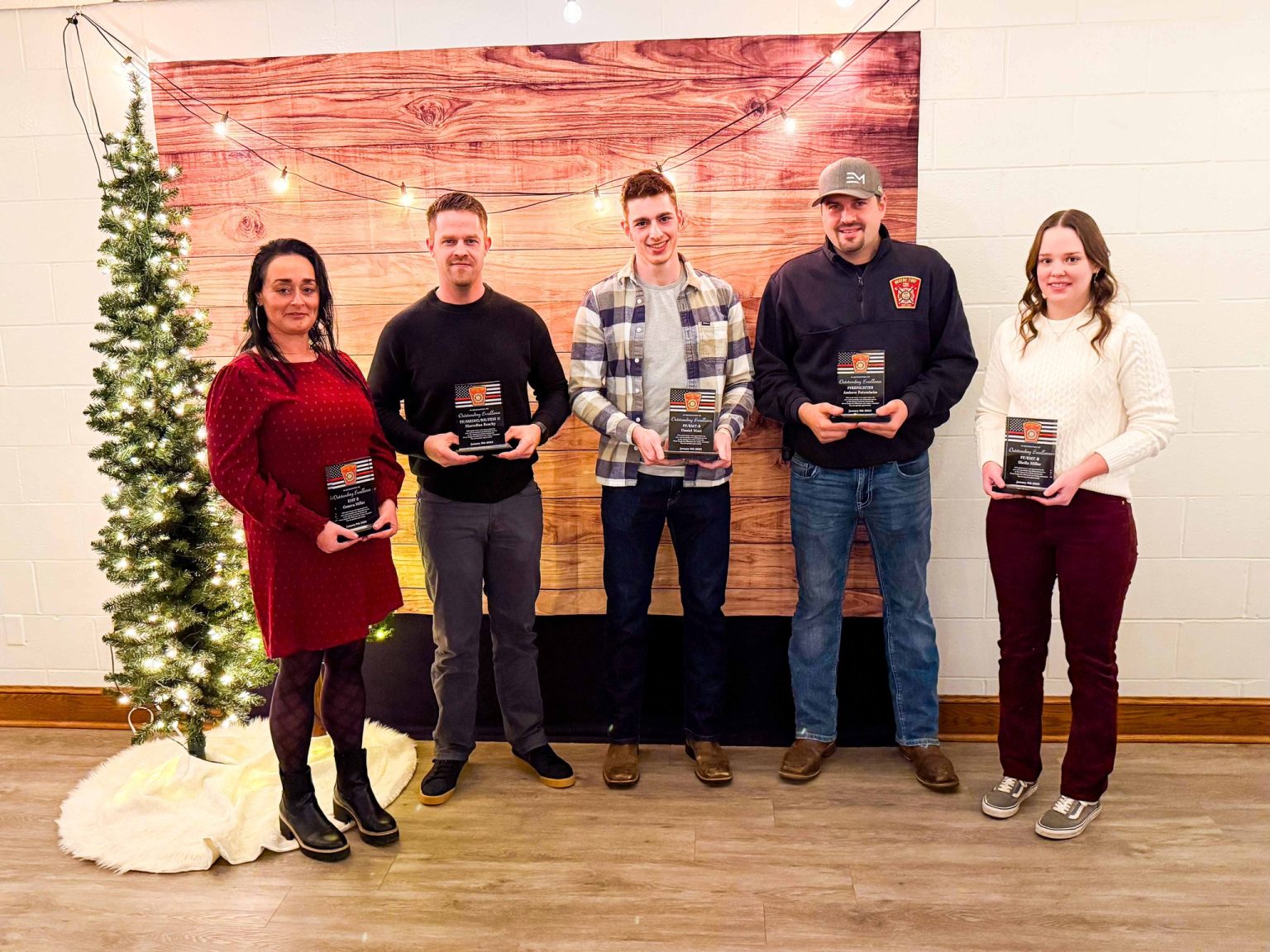 Five people stand in a row indoors, each holding a plaque. They are in front of a wooden backdrop and a decorated Christmas tree with lights. They are dressed casually and smiling.