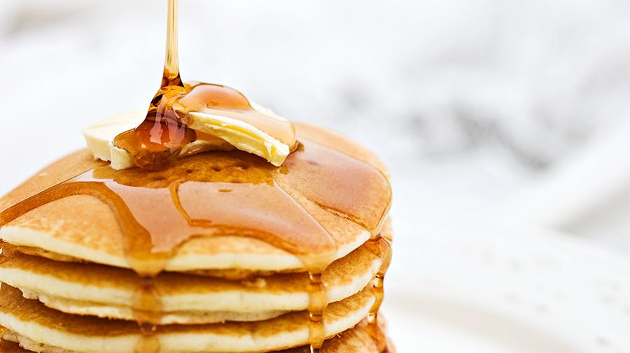 A stack of pancakes topped with a pat of butter being drizzled with syrup. The pancakes are on a white plate, and the syrup is flowing down the sides, creating a glossy surface. The background is softly blurred.