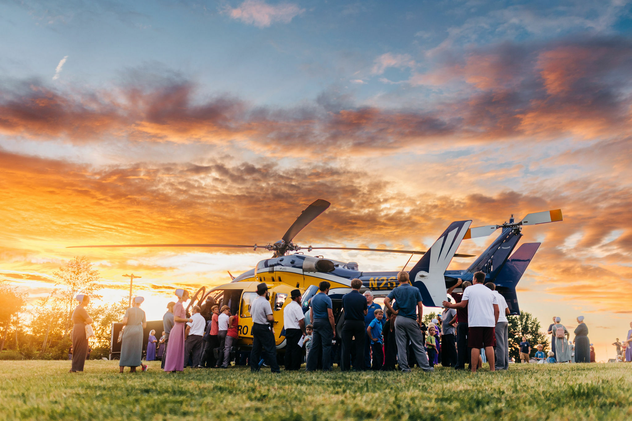 A large group of people gather around a yellow and blue helicopter on a grassy field. The sky is filled with dramatic orange and blue hues from a sunset.