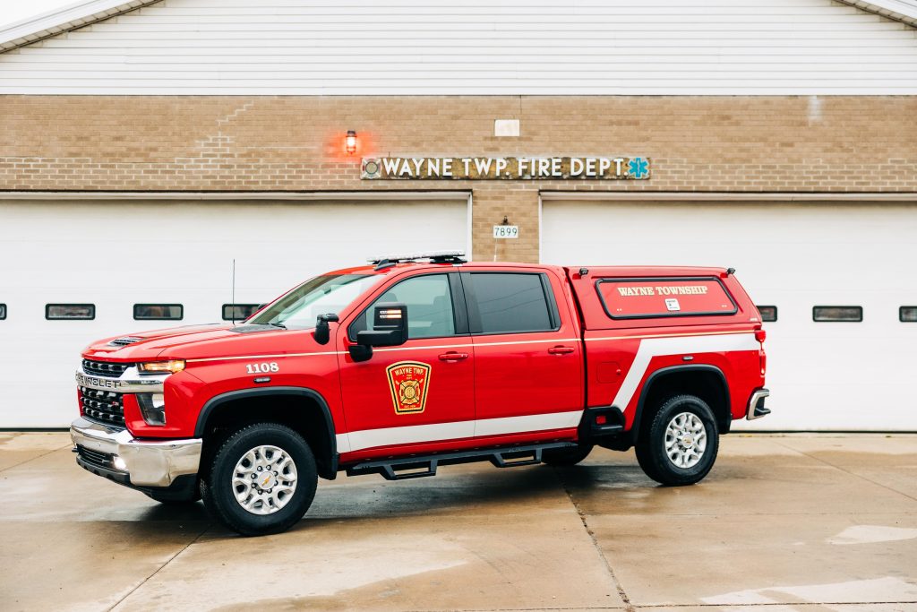 Red Wayne Township Fire Department truck parked in front of the fire station's garage doors. The truck is a Chevy with emergency symbols and department insignia. The station's building is beige brick with "Wayne Twp. Fire Dept" sign above.