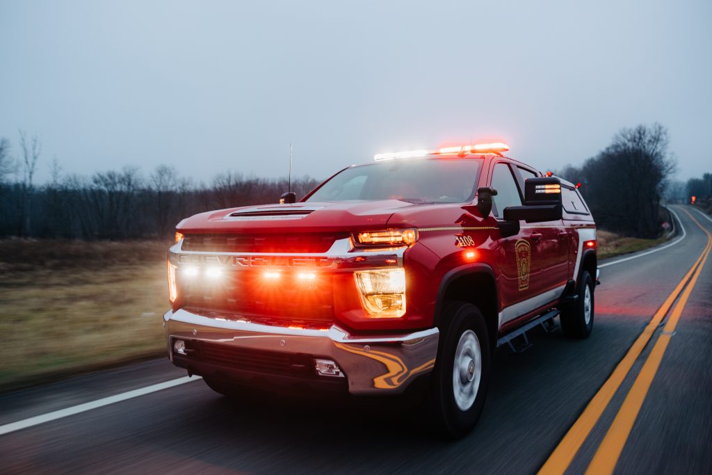 A red fire truck with flashing lights is driving on a rural road with a double yellow line. The surrounding landscape is overcast, with leafless trees and grassy fields visible in the background.