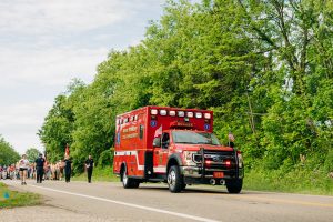 A red ambulance with American flags on the front drives down a tree-lined road. A group of people walks in the background on the left side of the road under a partly cloudy sky.