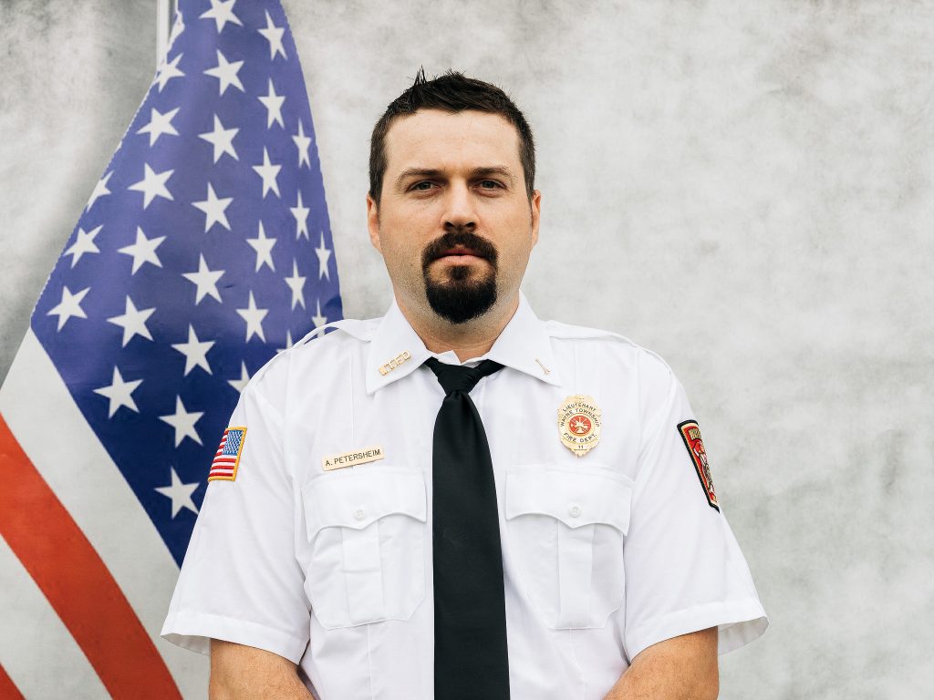 A man in a white uniform shirt with patches and a black tie stands in front of a U.S. flag. The background is a plain light gray wall.
