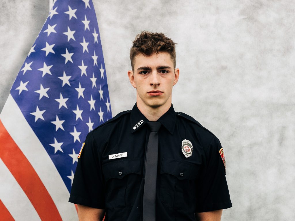 A young man in a dark uniform with a badge and name tag stands in front of an American flag. The name tag reads "D. Mast." The background is a neutral gray color.