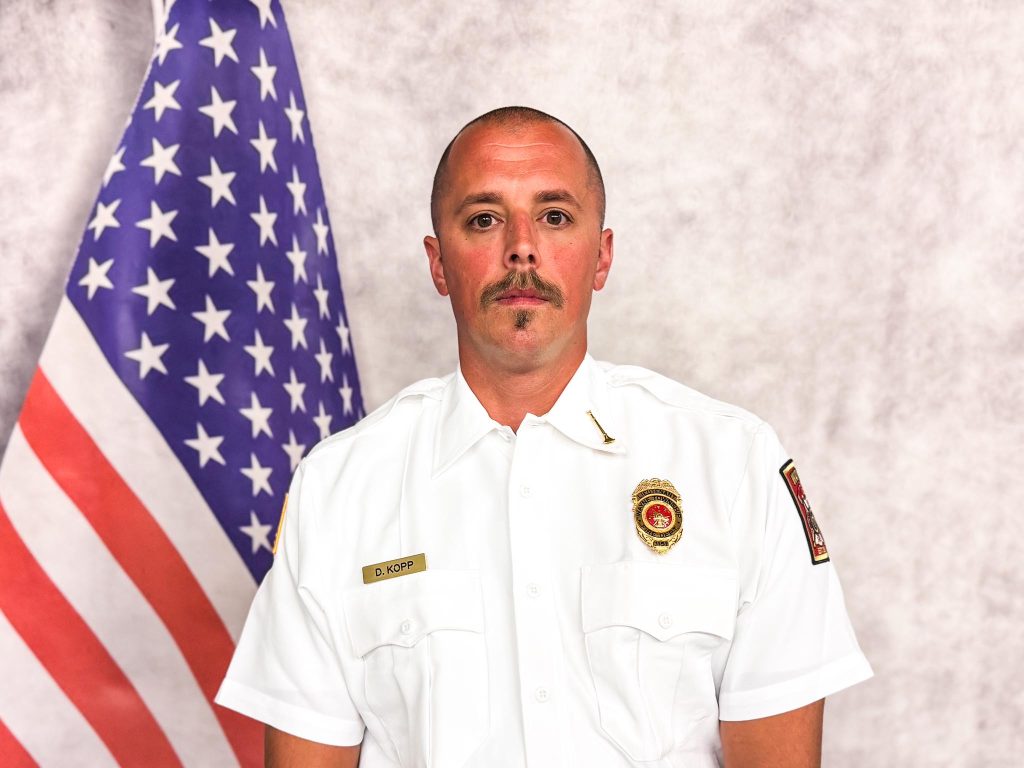 A firefighter in a white uniform with a badge and name tag stands in front of a U.S. flag and a gray backdrop, looking directly at the camera.