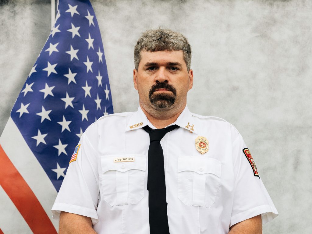 A firefighter in a white uniform with a patch that reads "FIRE DEPT" stands in front of an American flag. He has short hair and a mustache, and wears a black tie and various badges. The background is a plain, light-colored wall.