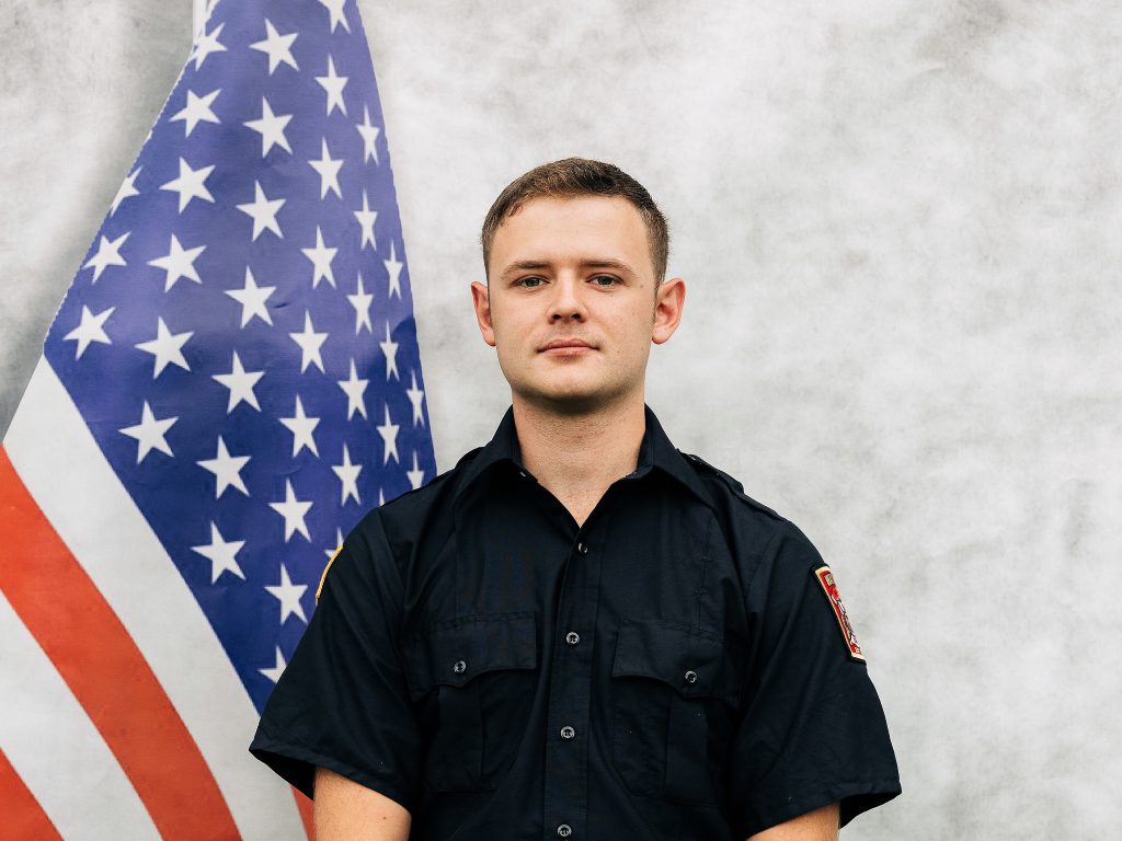 A person in a navy blue uniform stands in front of an American flag, set against a light gray background.