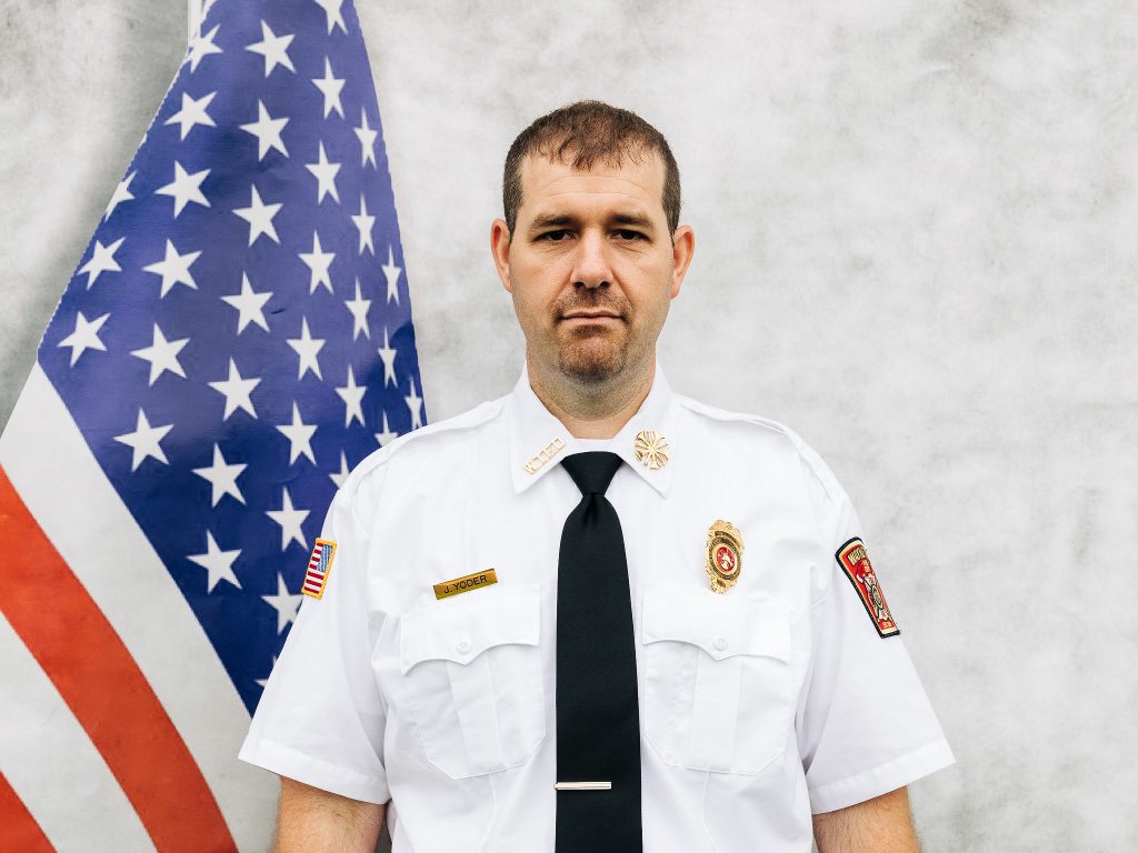 A man in a white uniform with patches and a black tie stands in front of an American flag, looking at the camera. The uniform has a badge and name tag. He has short hair and a neutral expression.