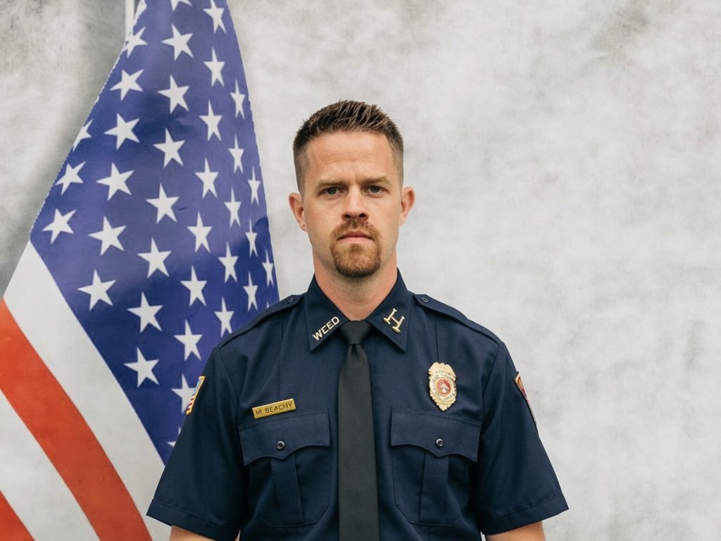 A uniformed firefighter stands in front of a U.S. flag and a light gray background, looking directly at the camera with a serious expression.