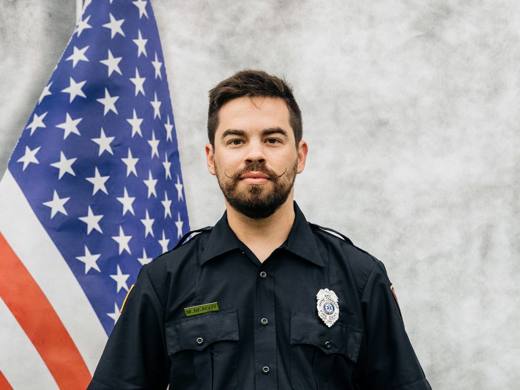 A man with a beard and styled mustache stands in front of an American flag. He is wearing a dark uniform with a badge and name tag. The background is a light, textured gray.