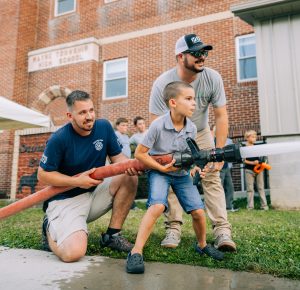 A young boy holds a large fire hose with assistance from two men, one kneeling and one standing. They are outside, near a brick building, on a grassy area with people in the background. The scene conveys teamwork and excitement.