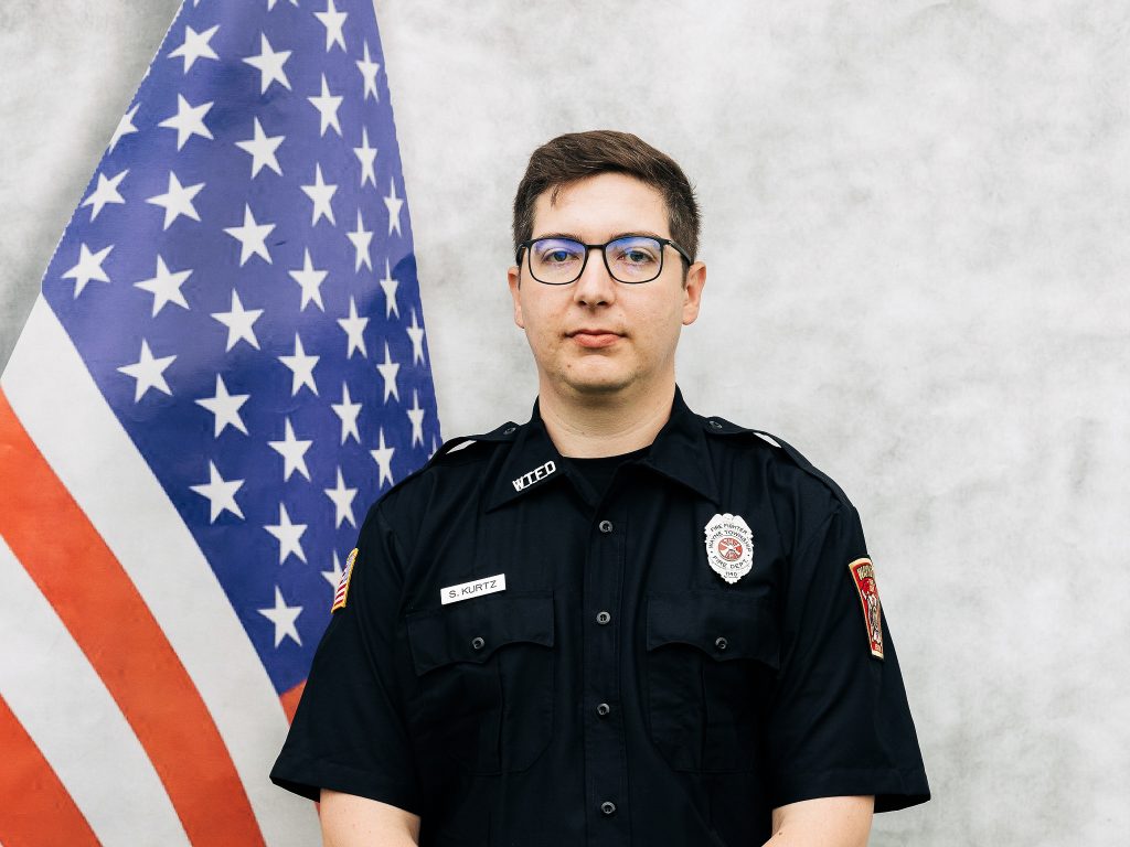 A uniformed officer stands in front of a U.S. flag. The officer, wearing glasses and a black uniform with a badge and name tag, poses against a neutral background.