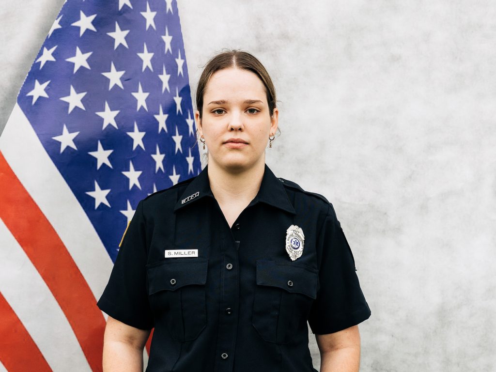 A person in a police uniform stands in front of an American flag. They have a neutral expression and are wearing a dark short-sleeved shirt with a badge and name tag. The background is a light gray color.