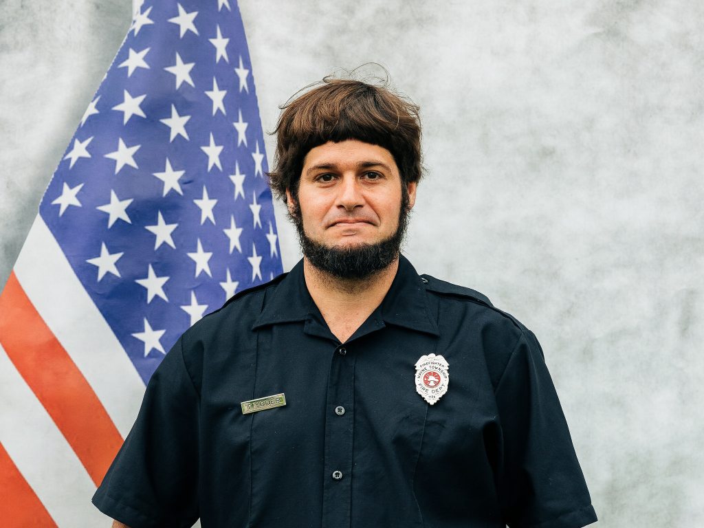 A person wearing a dark uniform with a badge stands in front of an American flag. The background is light gray.