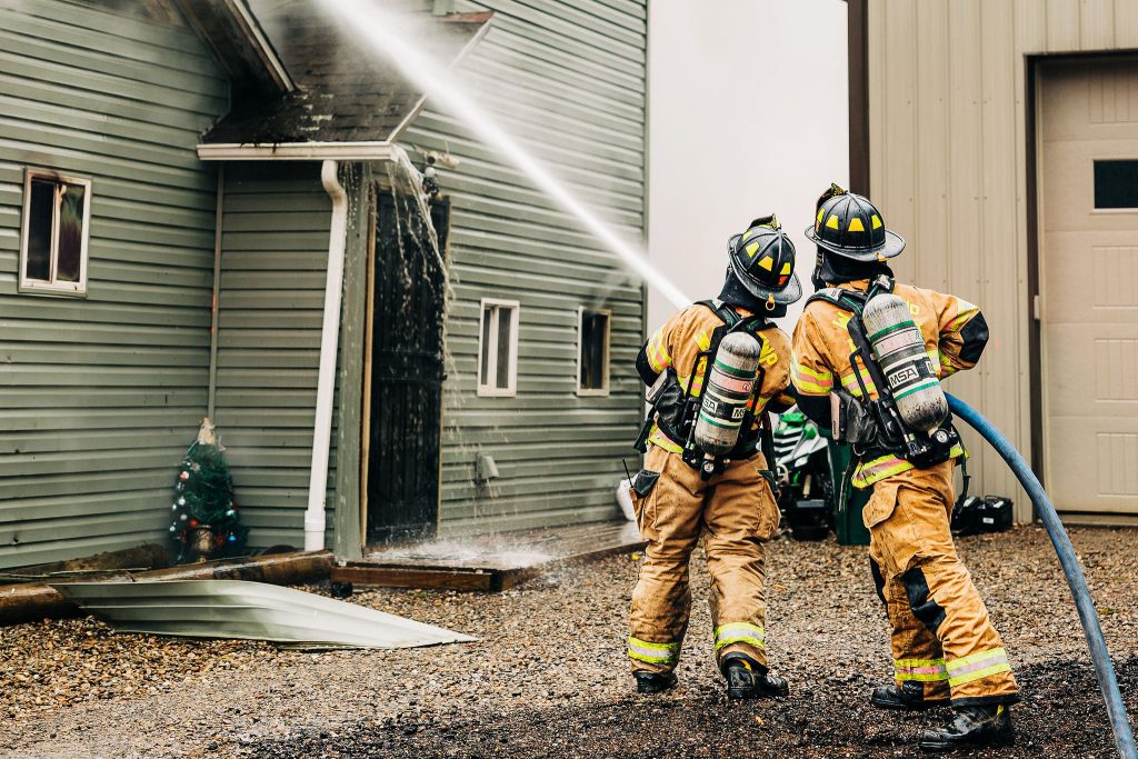 Two firefighters in yellow gear spray water on a two-story building using a hose. One holds the hose while the other directs the water stream towards the roof. Nearby, a metal sheet and a Christmas tree with lights are visible on the ground.